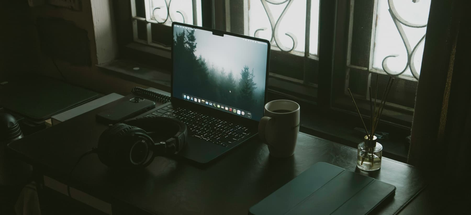 A laptop, headphones and a mug of coffee on a desk by a window