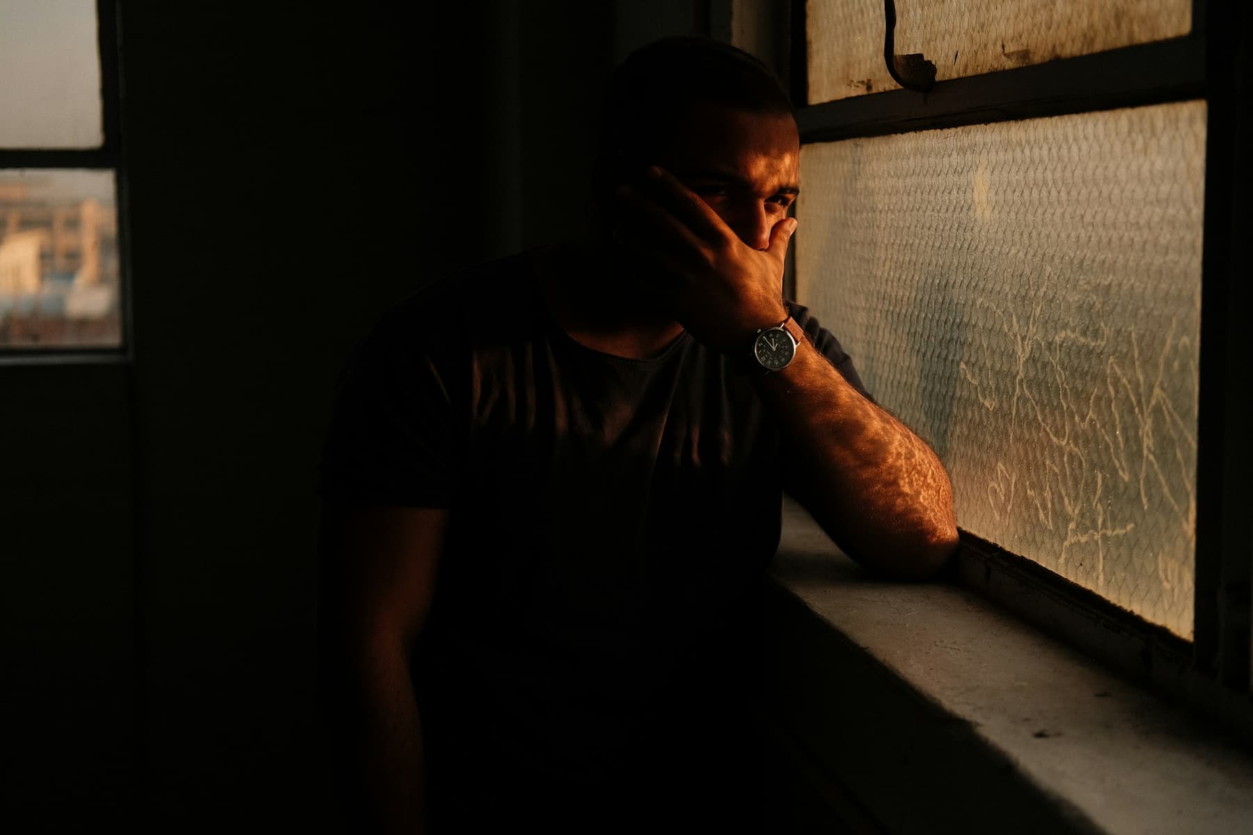 A man leaning by a window in a dark room, on the phone