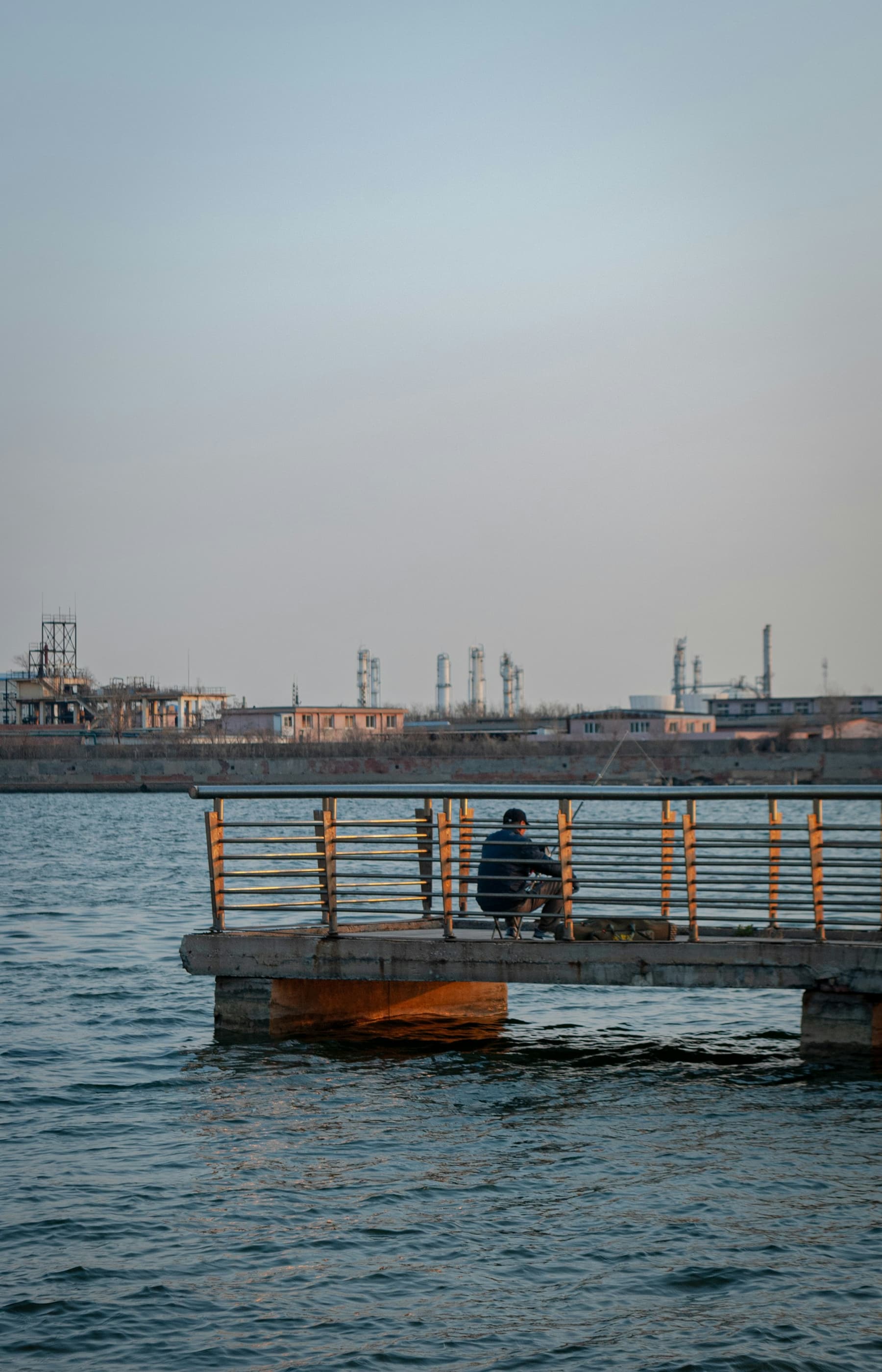 A man sitting at the end of a pier with an industrial waterfront behind