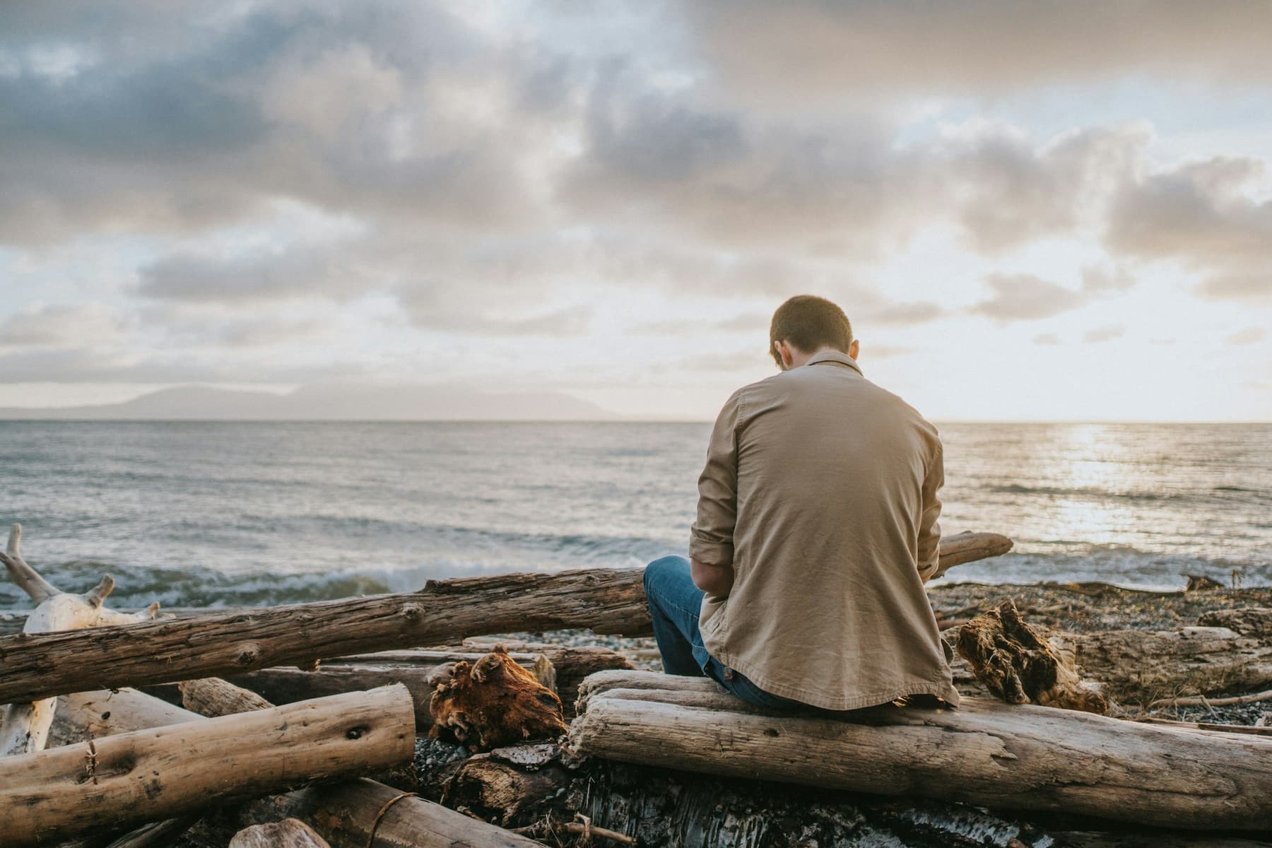 A man sitting on driftwood on a shoreline, looking out at the water